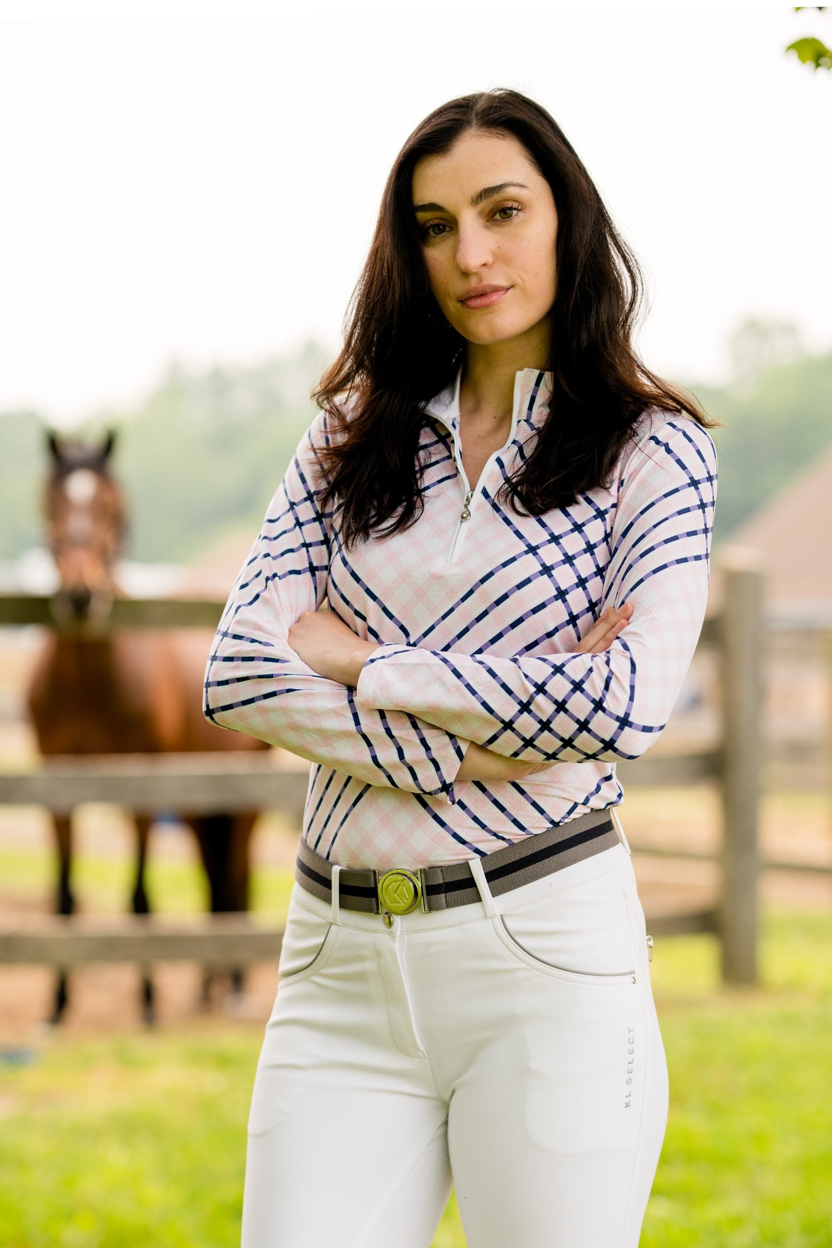 Front view of a female model wearing the white KL Select Gabrielle Breech with a horse standing in the background