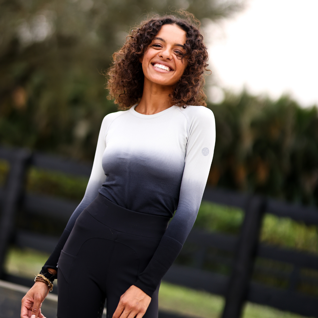 Front view of a female model wearing the Ombre TKEQ Kennedy Seamless Long Sleeve Shirt 2.0 while standing in front of a black wooden fence outside