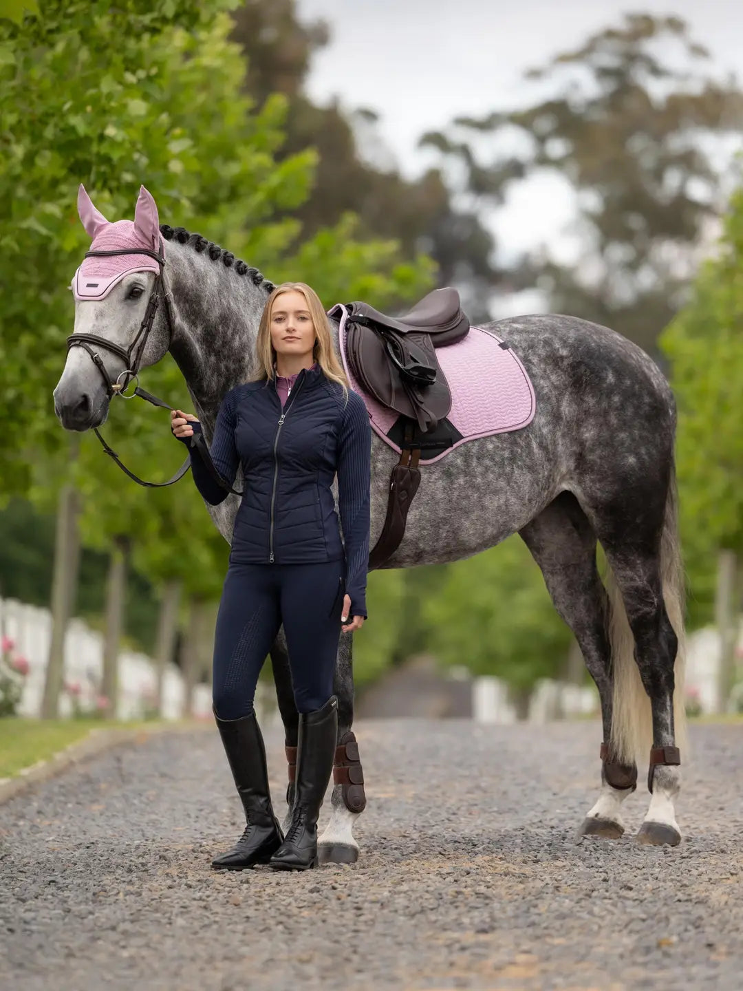 A woman standing outside in front of a gray horse with a LeMieux Essence Close Contact Square in pink under a brown saddle
