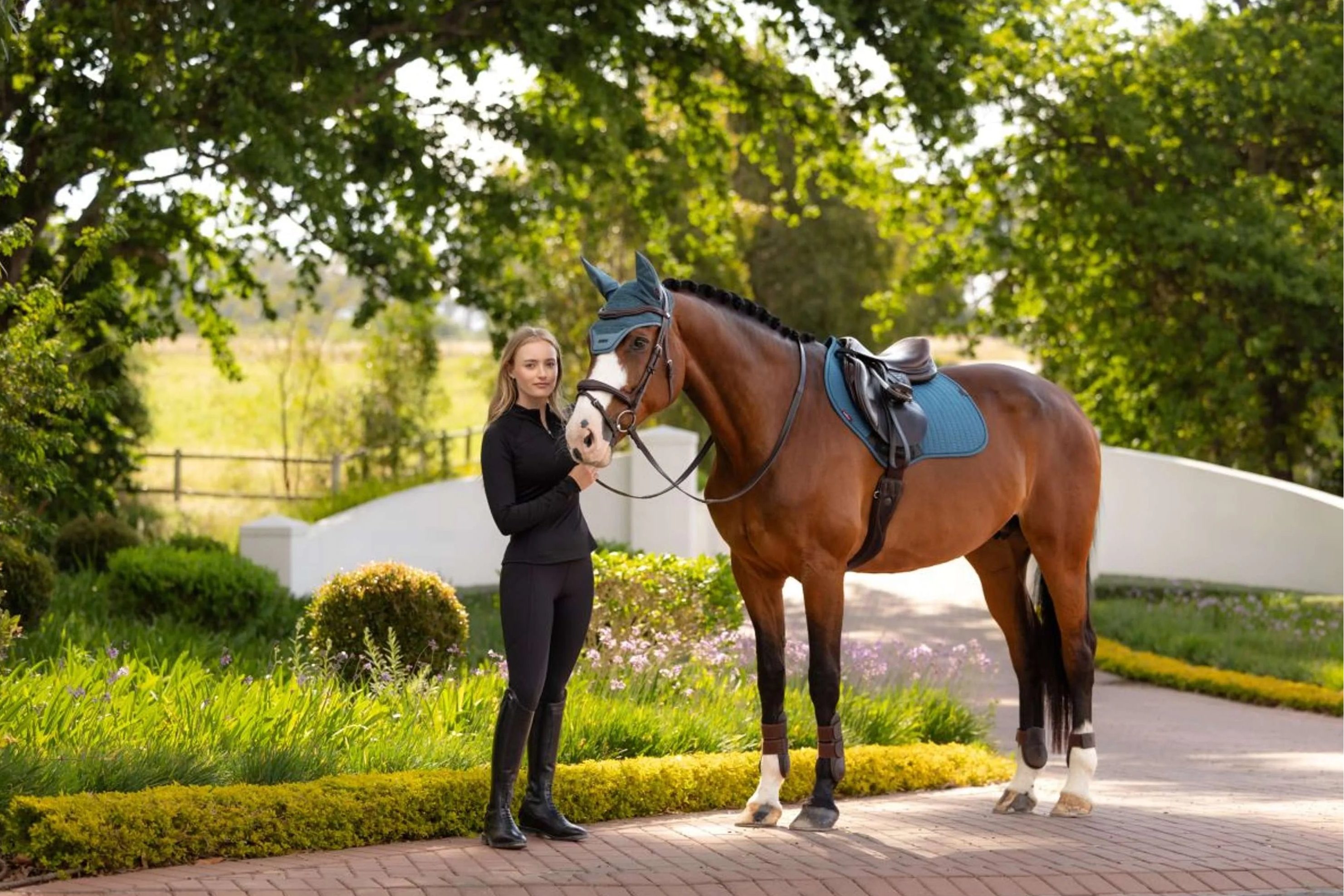 Woman standing next to a bay horse wearing the LeMieux Essence Close Contact Square in blue-green under a brown saddle