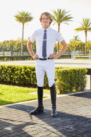 Man standing outside near and arena while wearing the white RJ Classics Tyler Show Shirt, navy tie, white breeches, and black tall boots