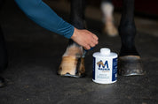 View of a person applying Mavrik Hoof Oil to a horse's front hooves with a 32 oz bottle on the ground