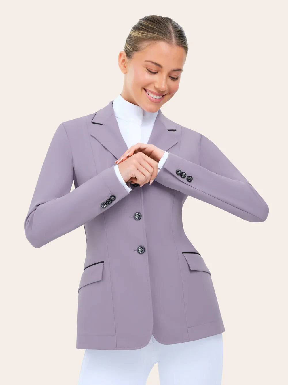 Angled front view of the RJ Classics Monterey Show Coat Eleganza Edition in Dusty Lilac, featuring black collar and pocket piping, three-button front closure, and logo buttons at the cuffs, shown over a white high-collar show shirt and white riding breeches on a model against a plain light background.