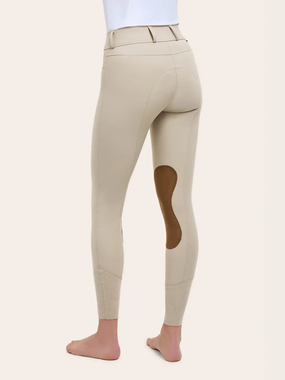 Angled back view of the RJ Classics Olivia Luxe-Tek Breech in Sand showing the contoured seat seams, belt loops, smooth stretch fabric, brown suede knee patch, and perforated lower-leg panels, photographed on a model against a plain light background.