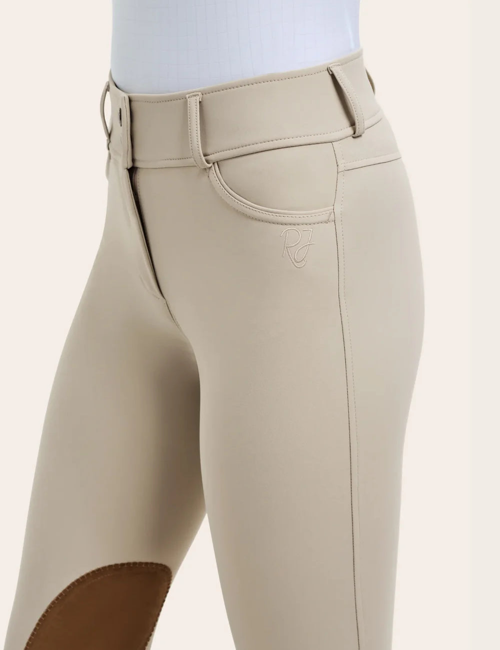 Close-up of the RJ Classics Olivia Luxe-Tek Breech in Sand showing the wide waistband with belt loops, zip front closure, front pocket, and RJ logo embroidery above the brown suede knee patch.