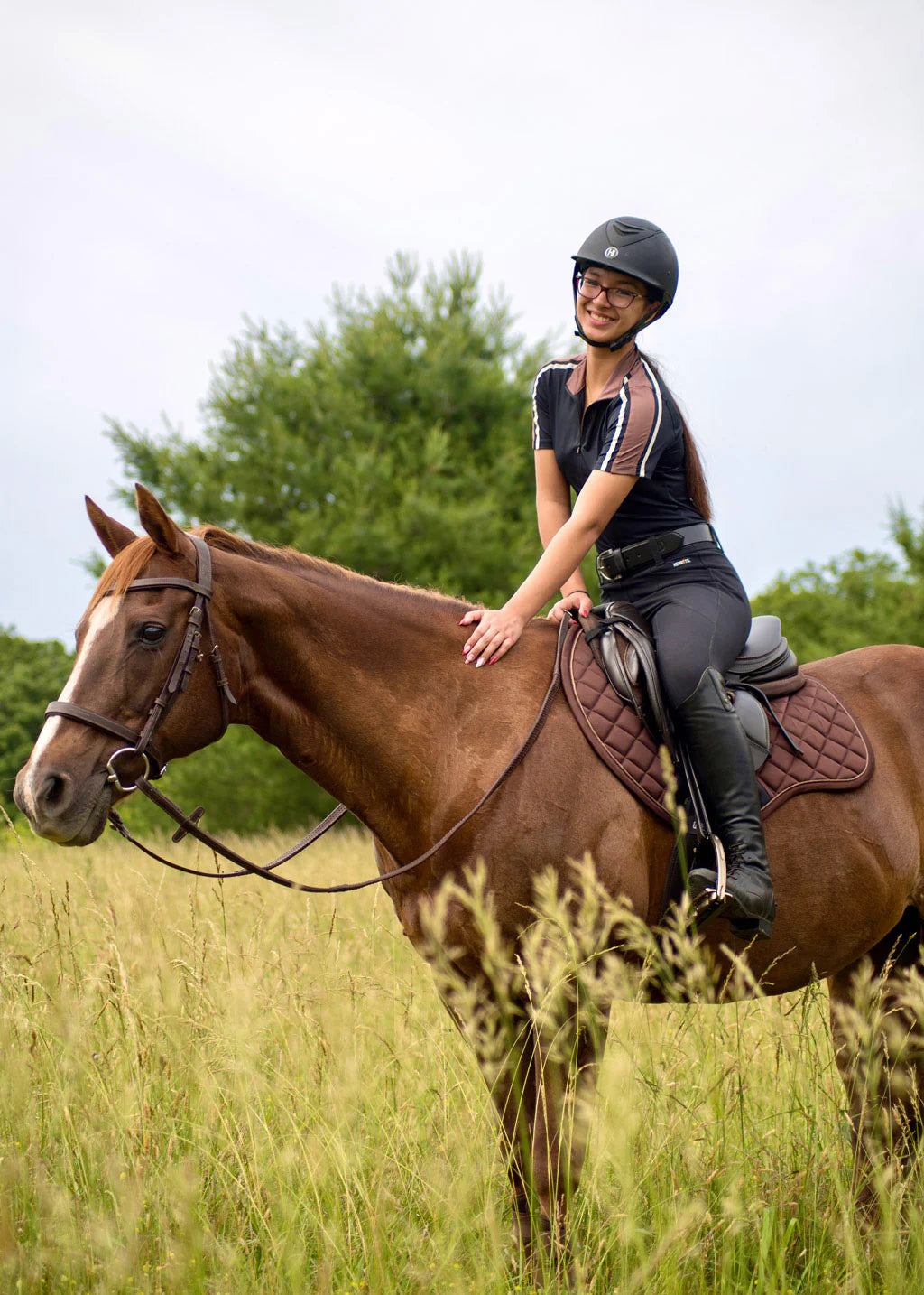 Side view of a female model riding a chestnut horse that is wearing the mocha Kerrits Close Contact Saddle Pad while standing in a field of tall grass