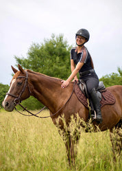 Side view of a female model riding a chestnut horse that is wearing the mocha Kerrits Close Contact Saddle Pad while standing in a field of tall grass