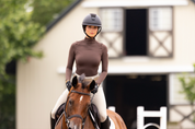 Front view of a female model wearing the Espresso TKEQ Chloe High Collar Technical Top while riding a horse