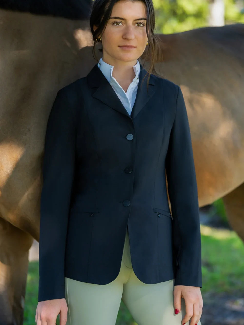 Lifestyle photo of the RJ Classics Sloane Show Coat in black, featuring a notched lapel, three-button front closure, and slanted zip pockets, worn over a white collared shirt with tan breeches beside a horse outdoors.