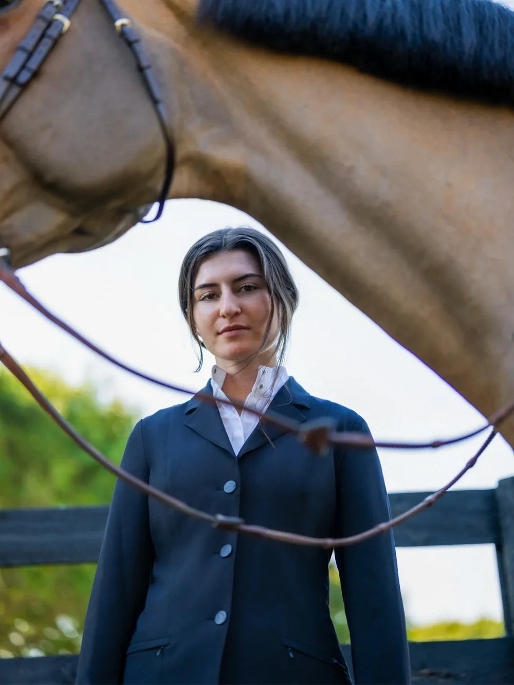 Lifestyle photo of the RJ Classics Sloane Show Coat in black with a three-button front and slanted zip pockets, worn over a white collared shirt, shown outdoors with a horse overhead and reins in the foreground.