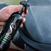 View of someone spraying the 16oz Eucalyptus Sterling Essentials Leather Cleaner on a black saddle