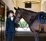 Front view of a woman wearing the navy Aubrion Optima LX Show Coat  while standing in a stable next to a dark colored horse