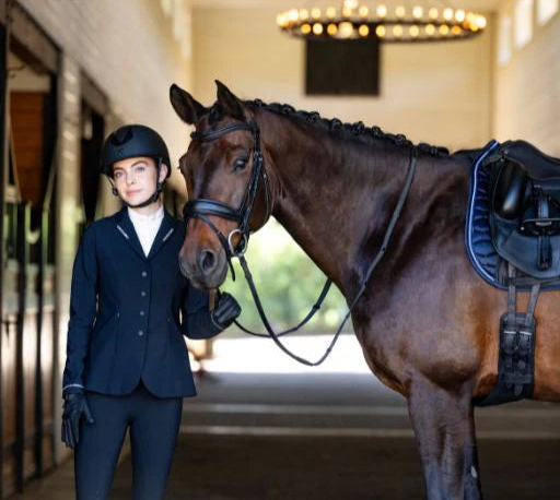 Front view of a woman wearing the navy Aubrion Optima LX Show Coat  while standing in a stable next to a dark colored horse