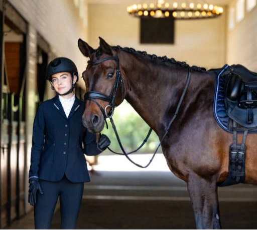 Front view of a woman wearing the navy Aubrion Optima LX Show Coat  while standing in a stable next to a dark colored horse