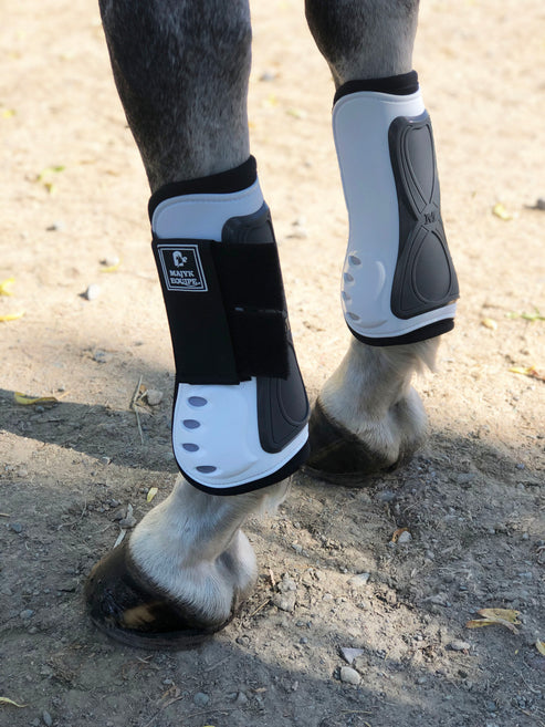 Angled back view of a gray horse wearing the white Majyk Equipe Vented Infinity Open Front Tendon Jump Boot while standing in the dirt outside