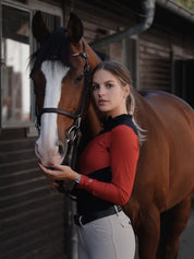 Front view of a bay horse and a female model who is wearing the black/red Criniere Sophie Fierce Focus Long Sleeve Shirt with a barn in the background
