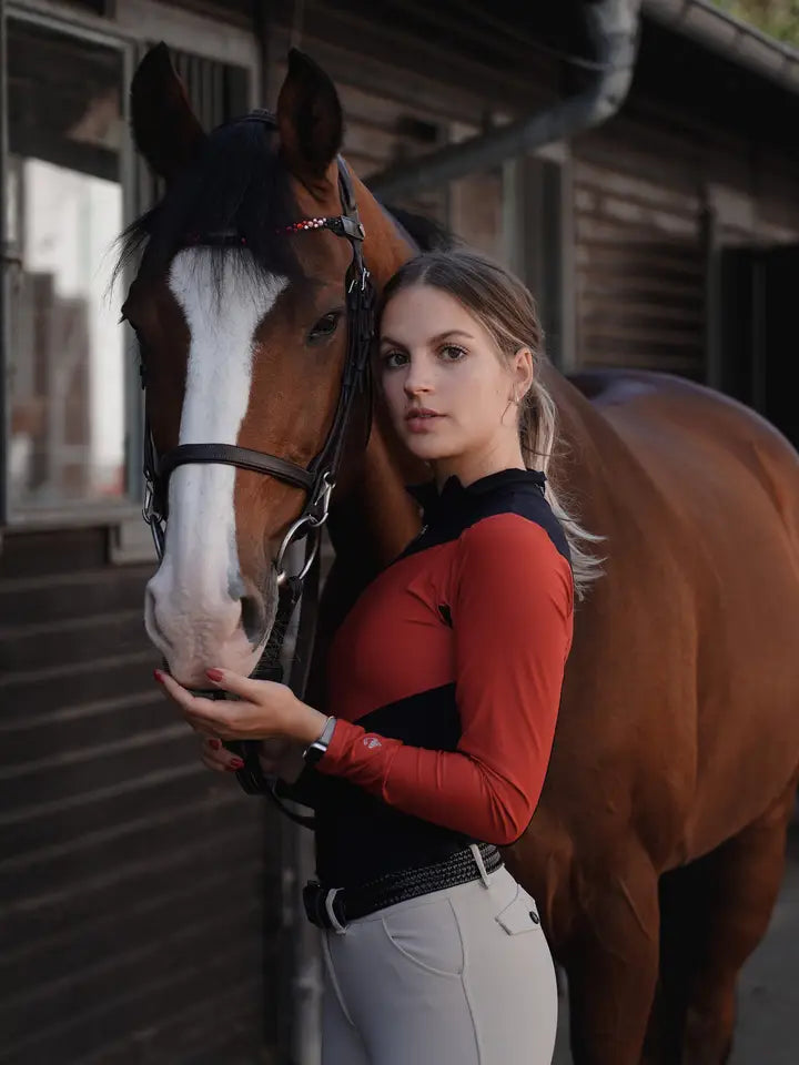 Front view of a bay horse and a female model who is wearing the black/red Criniere Sophie Fierce Focus Long Sleeve Shirt with a barn in the background