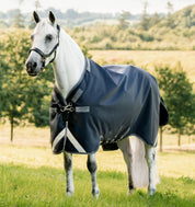 Angled front view of a white horse standing in a pasture while wearing the navy Horseware Rambo 1680D Turnout 100g