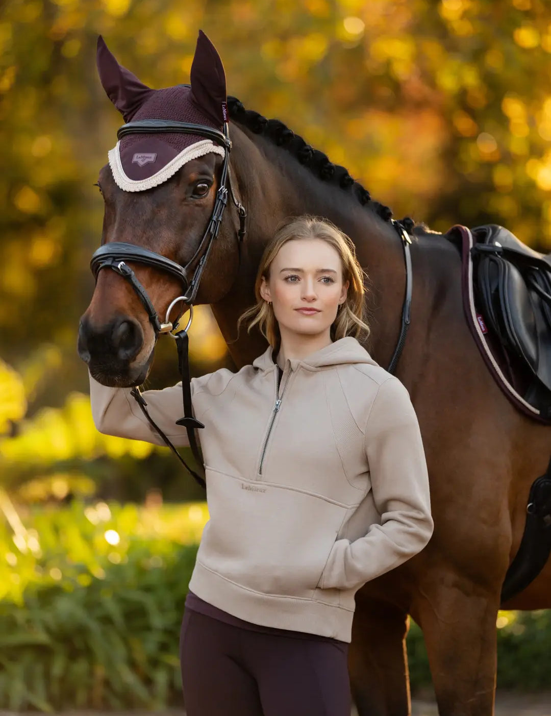 Front view of a model standing outside in front of a dark colored horse that is wearing the damson LeMieux Loire Fly Hood