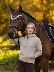 Front view of a model standing outside in front of a dark colored horse that is wearing the damson LeMieux Loire Fly Hood