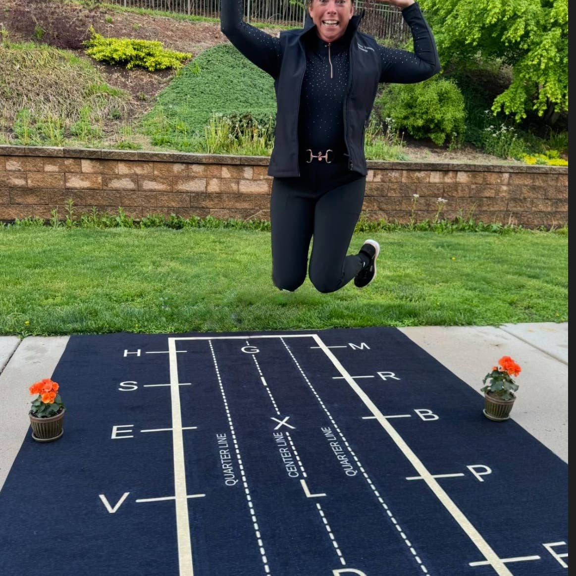 View of a model jumping on the All Designs Equine Dressage Arena Practice Your Test Outdoor Horse Trainer Rug on an outdoor patio