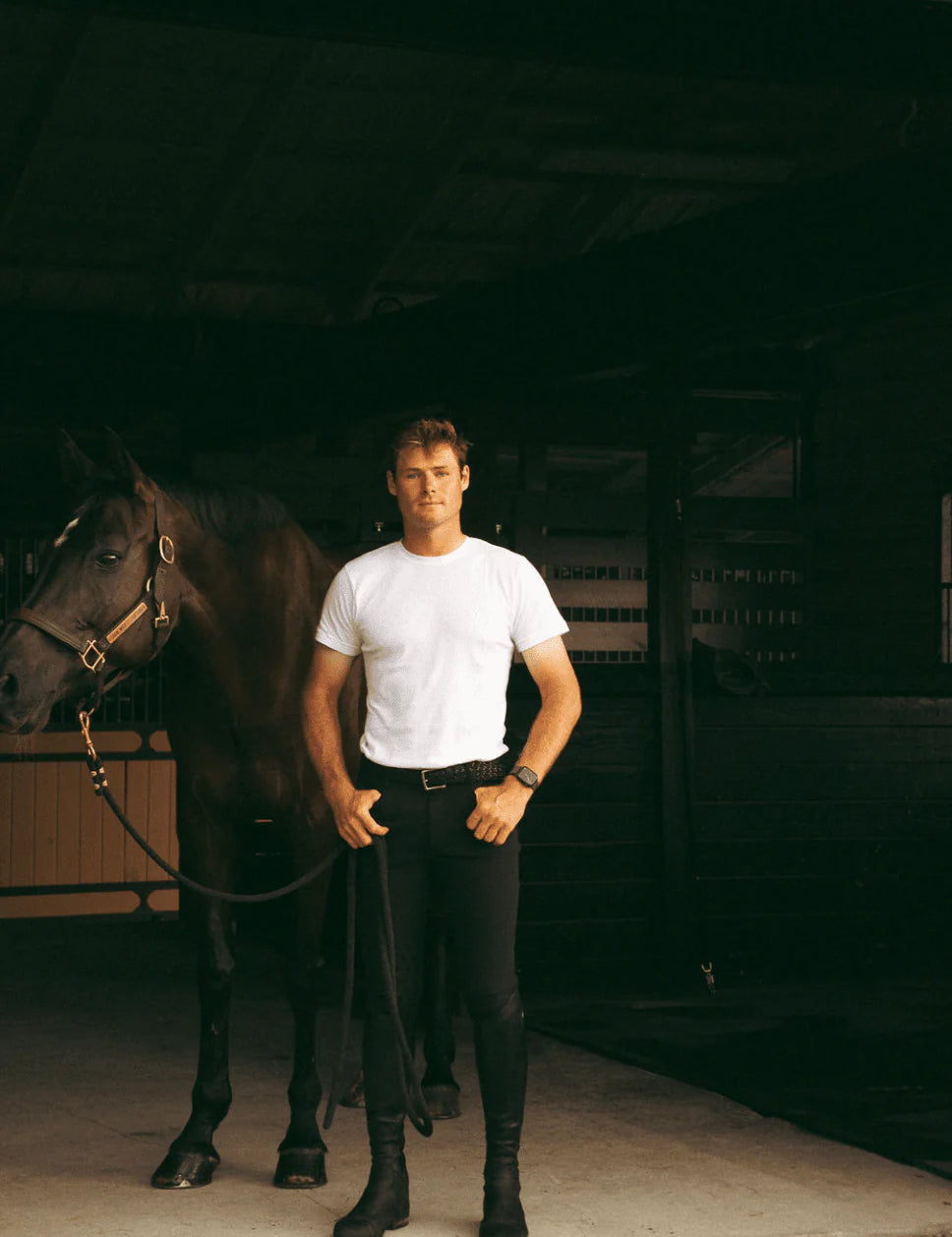 Full body front view of a male model wearing the black Criniere Men's Chadai Half Seat Breeches and white shirt while standing in front of a horse in a barn
