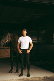 Full body front view of a male model wearing the black Criniere Men's Chadai Half Seat Breeches and white shirt while standing in front of a horse in a barn