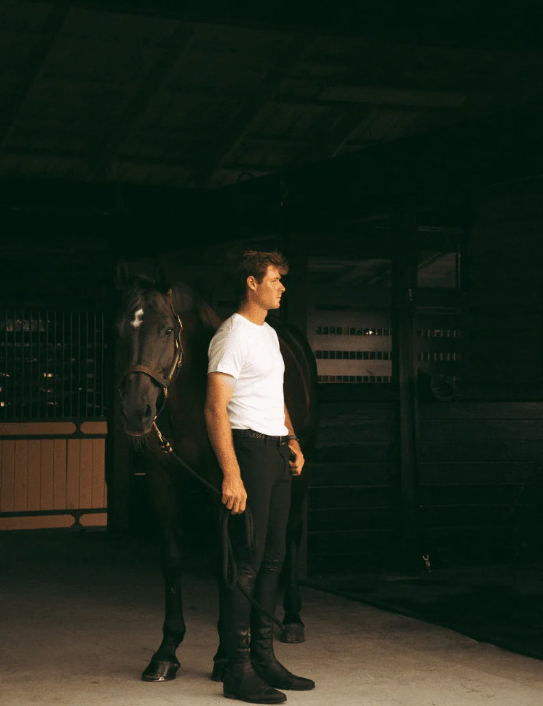 Full body angled front view of a male model wearing the black Criniere Men's Chadai Half Seat Breeches and white shirt while standing in front of a horse in a barn