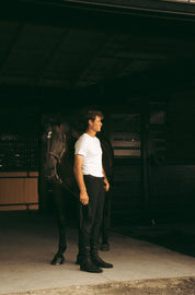 Full body angled front view of a male model wearing the black Criniere Men's Chadai Half Seat Breeches and white shirt while standing in front of a horse in a barn