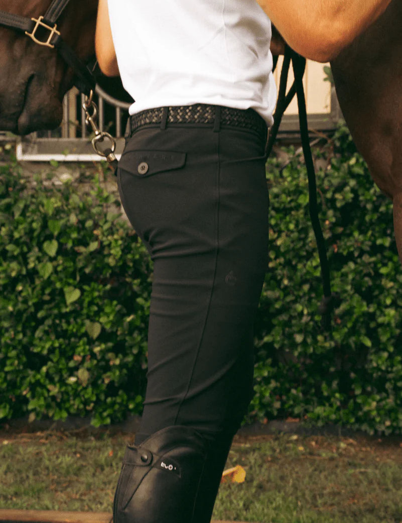 Side view of a model wearing the black Criniere Men's Chadai Half Seat Breeches and white shirt while standing in front of a horse outside