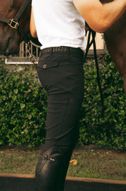 Side view of a model wearing the black Criniere Men's Chadai Half Seat Breeches and white shirt while standing in front of a horse outside