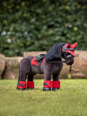 Angled front view of a dark colored toy pony wearing the chilli red LeMieux Toy Pony Fly Hood and matching set while standing in the grass