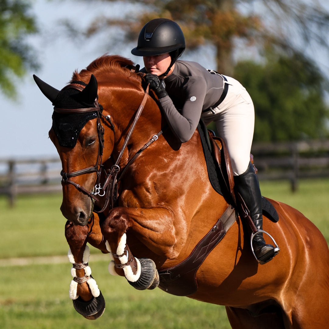 Angled front view of a female model wearing the Slate TKEQ Chloe High Collar Technical Top while riding a horse over a jump