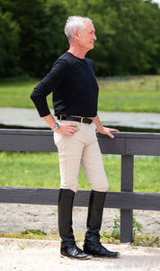 Side view of a male model wearing the beige KL Select Men's Evan Knee Patch Breeches while leaning up against a fence