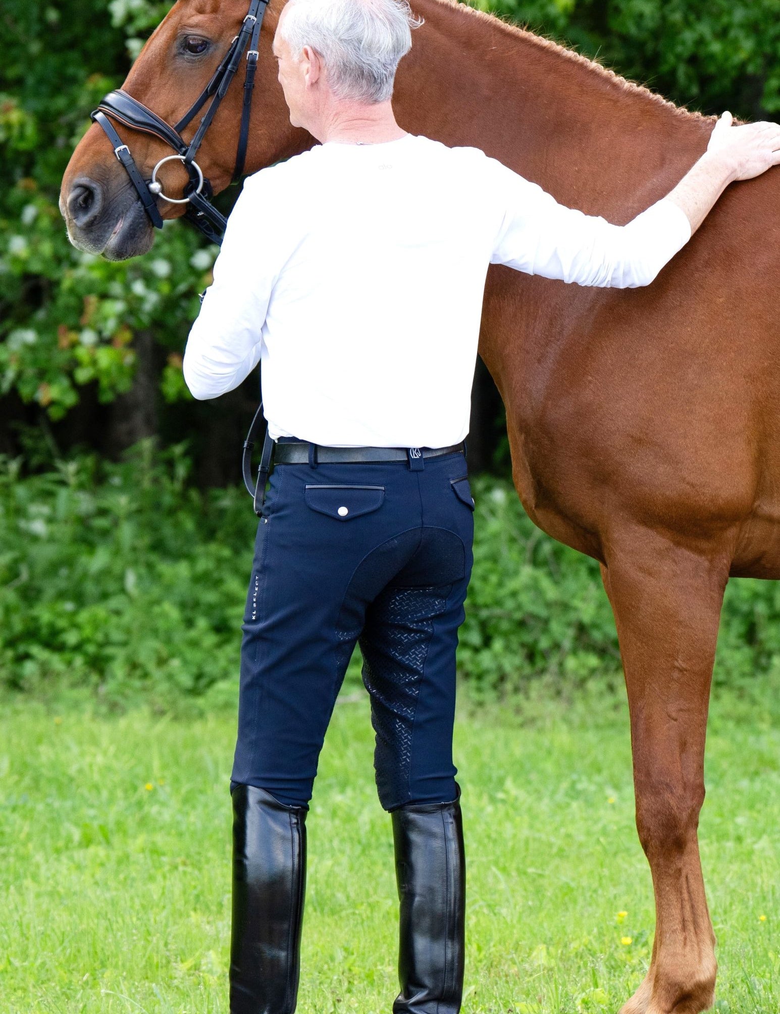 Back view of a male model wearing the navy KL Select Men's Evan Full Seat Breeches while petting a chestnut horse outside