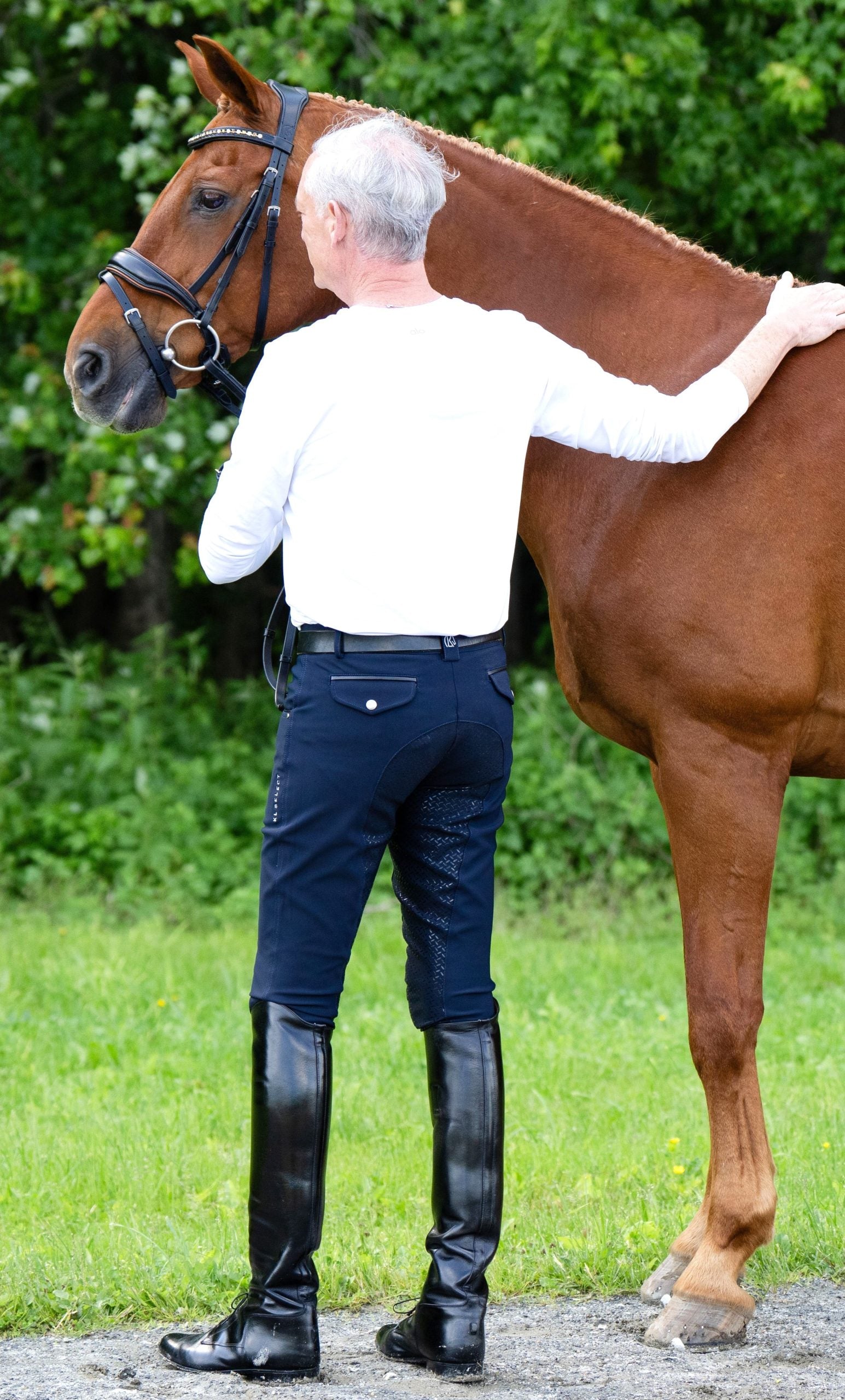Back view of a male model wearing the navy KL Select Men's Evan Full Seat Breeches while petting a chestnut horse outside