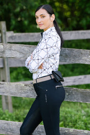 Side view of a female model wearing the black KL Select Gabrielle Full Seat Breech while standing outside in front of a wood fence