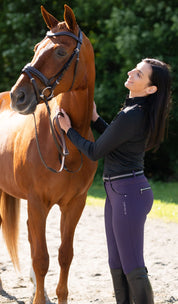 Side view of a model wearing the purple KL Select Gabrielle Breech while petting a chestnut horse