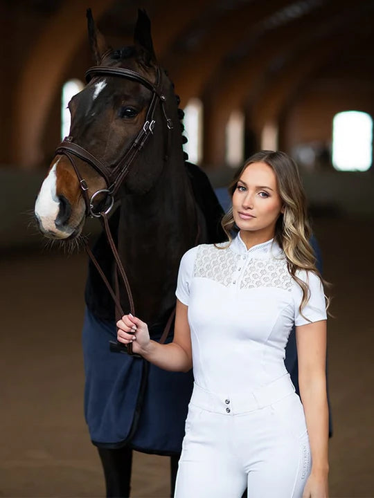 Front view of a female model wearing the white Equestrian Stockholm Crystal Champion Competition Top while standing nex to to a dark colored horse