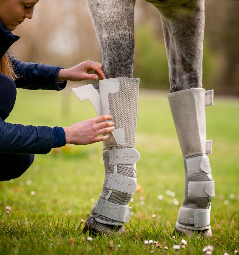 Person fastening Rambo Tech-Fit Fly Boot in silver on horse leg in grassy field