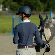 Back view of a woman wearing a blue top, tan breeches, and the navy brown-leather Equinavia Siri Polo Belt white leading a gray horse in an outdoor arena