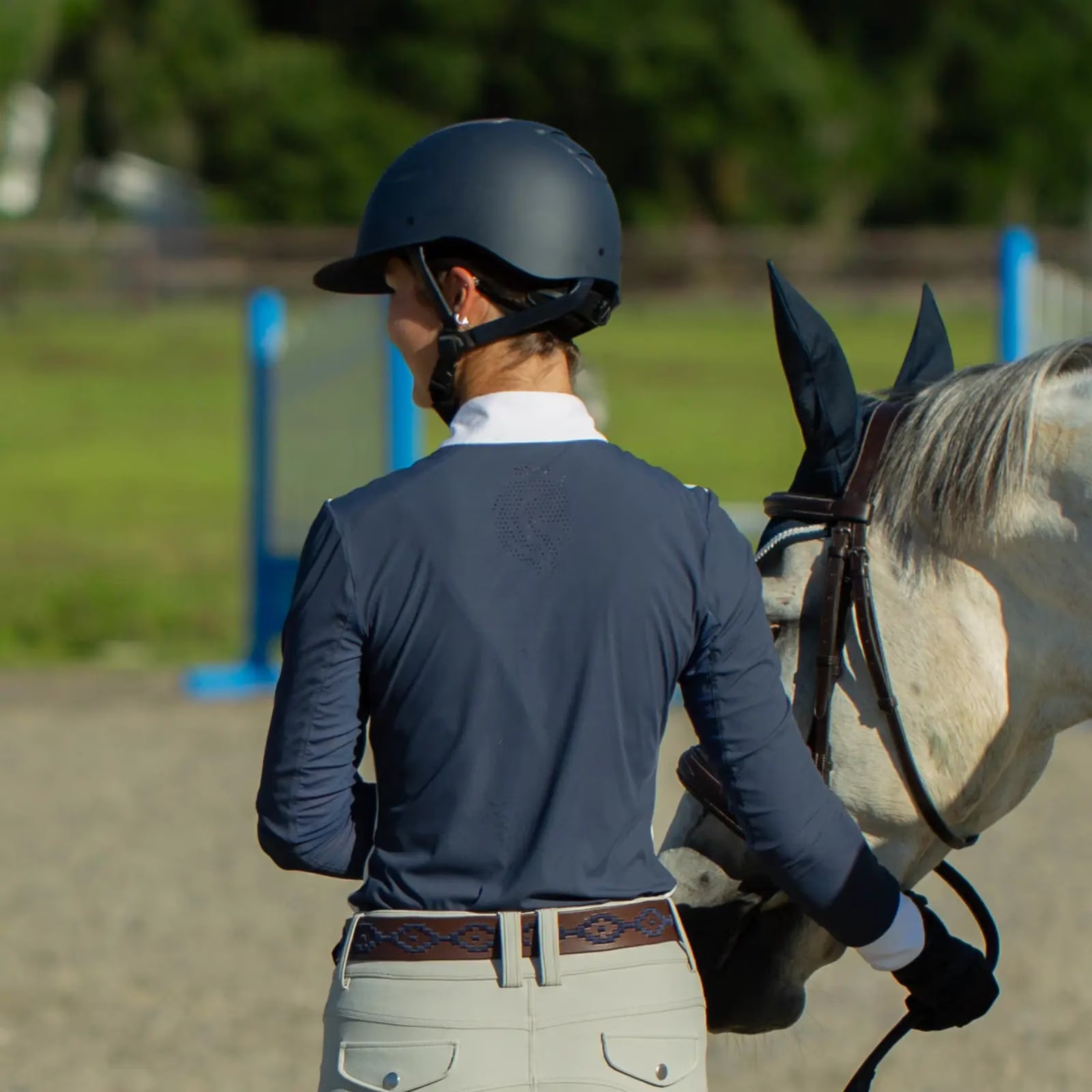 Back view of a woman wearing a blue top, tan breeches, and the navy brown-leather Equinavia Siri Polo Belt white leading a gray horse in an outdoor arena