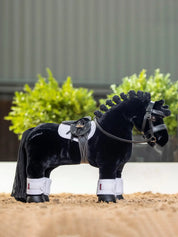 Side view of a black toy pony wearing the white LeMieux Toy Pony Saddle Pad while standing in an arena