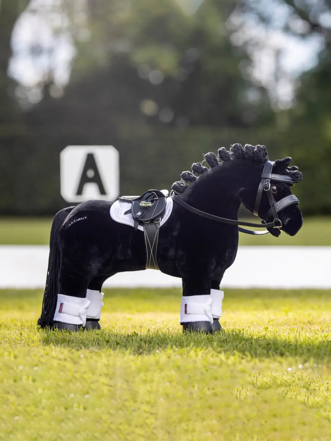 Side view of a black toy pony standing in a grass arena wearing the black LeMieux Toy Pony Saddle and Girth and white accessories