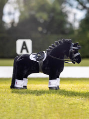 Side view of a black toy pony standing in a grass arena wearing the black LeMieux Toy Pony Saddle and Girth and white accessories