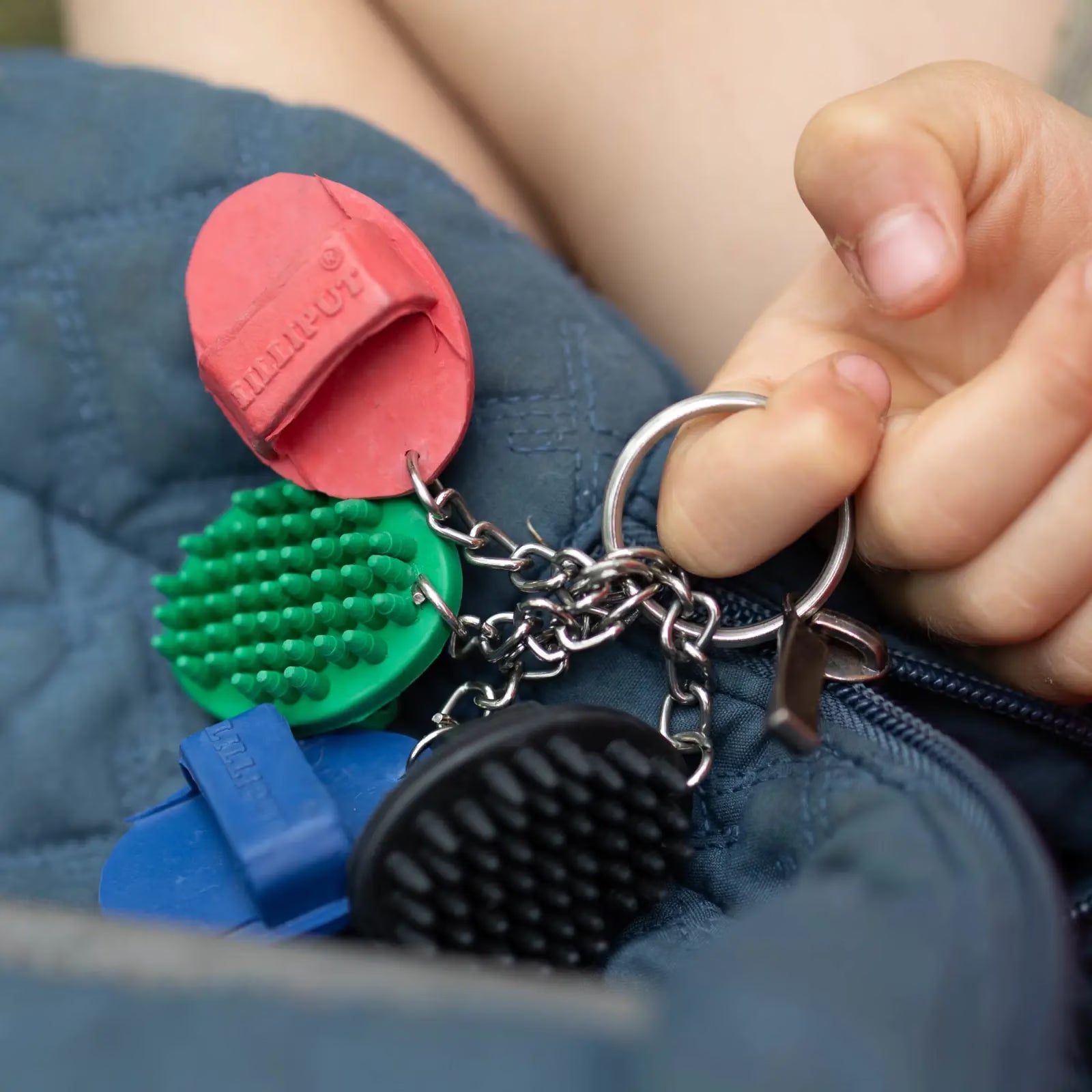 View of all the colors of the Horze Brush Key Chain (black, blue, green, red) on a blue zipper