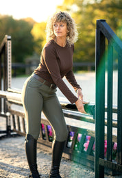 Angled front view of a female model wearing the olive green KL Select Gabrielle Full Seat Breech while standing in an outdoor jump arena
