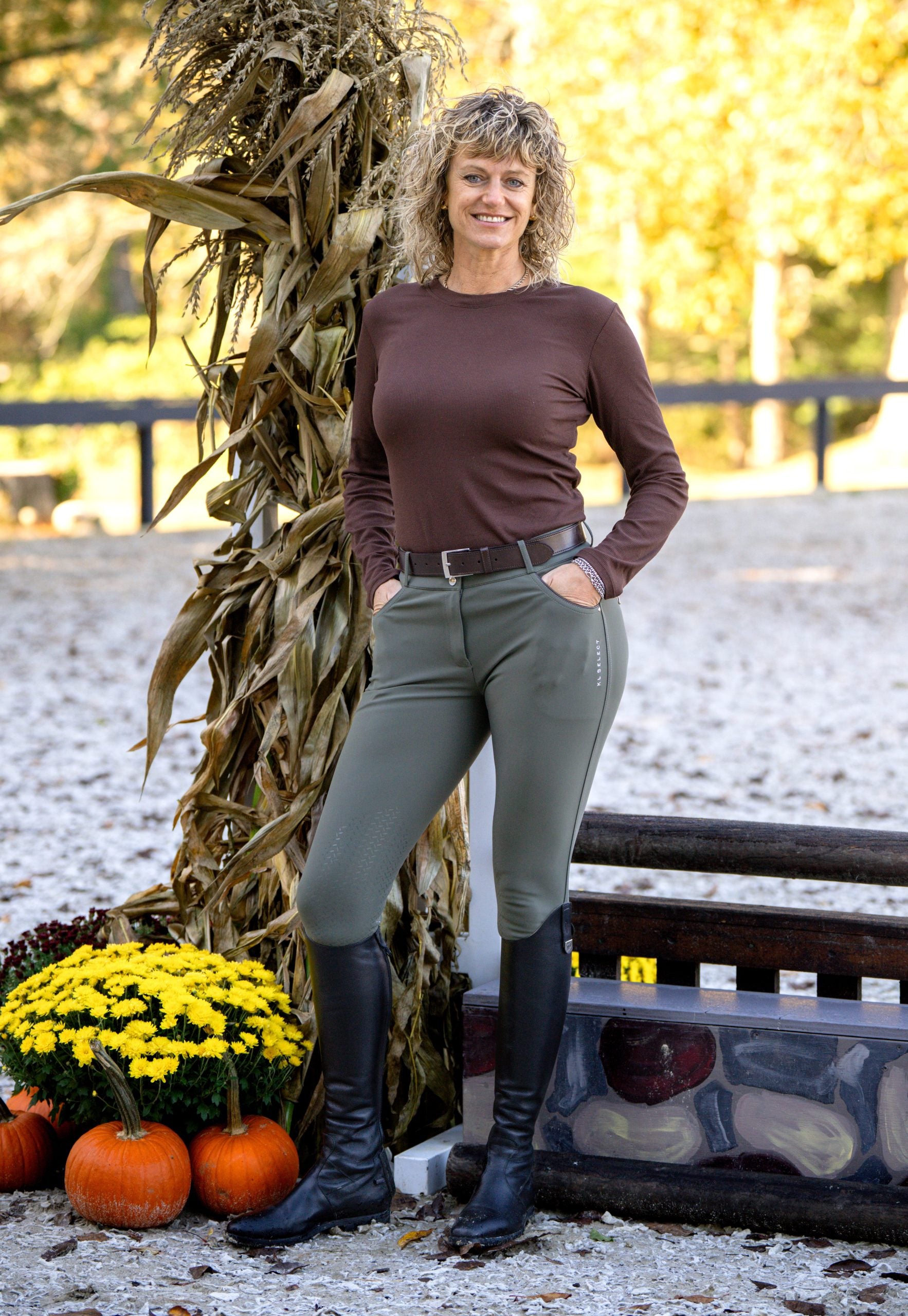 Full body front view of a female model wearing the olive green KL Select Gabrielle Knee Patch Breech while standing in an outdoor jump arena