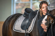 Angled side view of a dark colored horse wearing the Pale Grey Eskadron Mattgloss Contrast Jewel Dressage Saddle Pad under a black saddle with a female model standing in the foreground