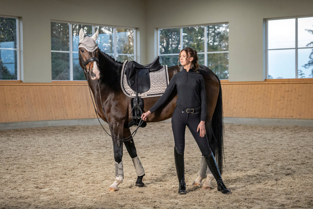 Side view of a dark colored horse wearing the Pale Grey Eskadron Mattgloss Contrast Jewel Dressage Saddle Pad while standing behind a female model in an indoor arena